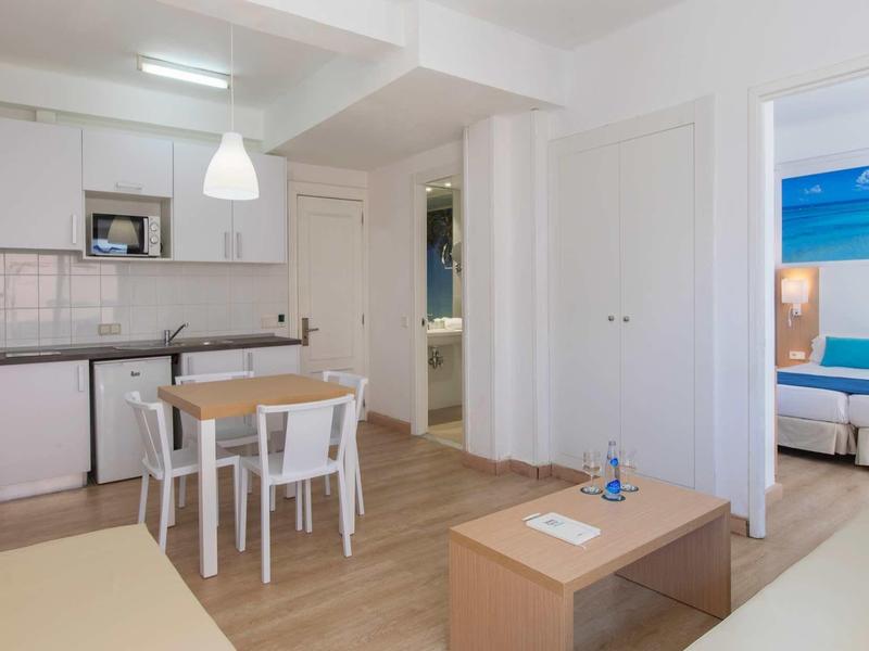 Modern living area with kitchen, dining table, and view into bedroom with blue headboard.