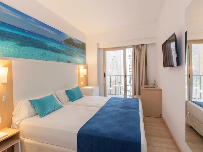 Bright hotel room with double bed, blue bedding, wall art, and glass door balcony.