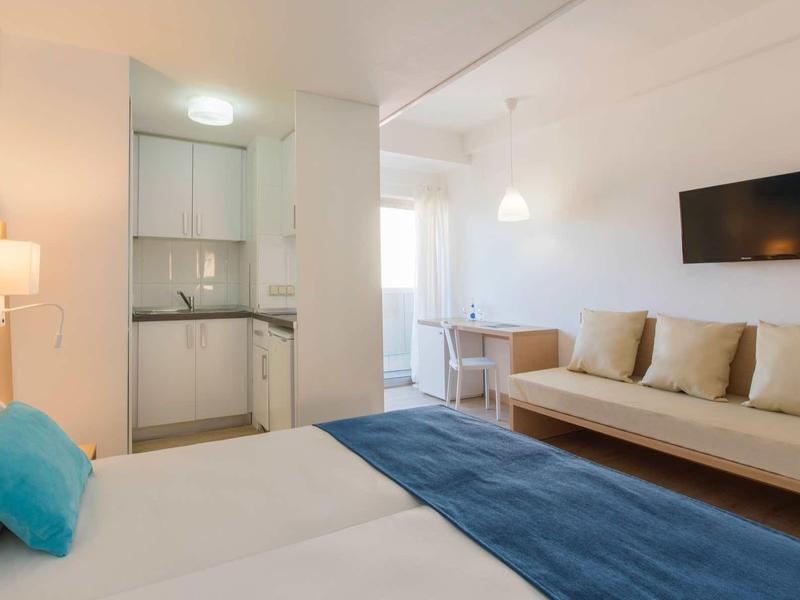 Bright hotel room with double bed, sofa, wall-mounted TV, and small dining table.