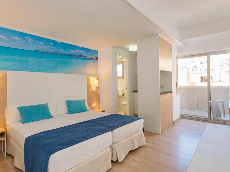 Bright hotel room with double bed, blue bedspread, ocean picture, and balcony.