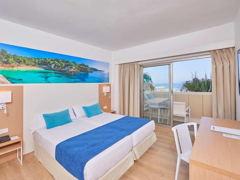 Bright hotel room with double bed, blue and white bedding, and balcony view of the sea.