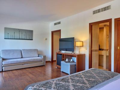 Hotelzimmer mit Bett, Sofa, TV und Holzmöbeln in warmem Licht.