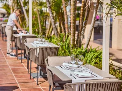 Außenterrasse mit gedeckten Tischen und einem Kellner neben Palmen.