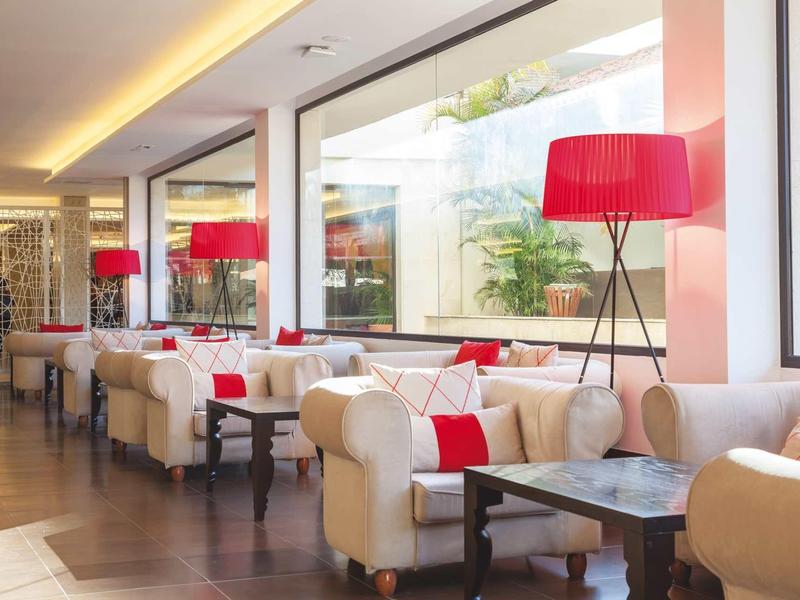 Area soggiorno luminosa dell'hotel con divani bianchi, cuscini rossi e lampade da terra moderne accanto a grandi finestre.