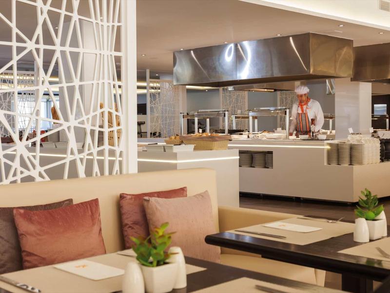 Lounge d'hotel moderna con posti a sedere, cuscini e cucina a vista sullo sfondo.