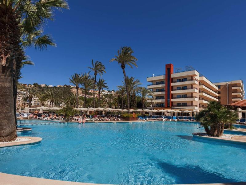 Grande piscina dell'hotel con palme e edificio su più piani sotto un cielo azzurro limpido.