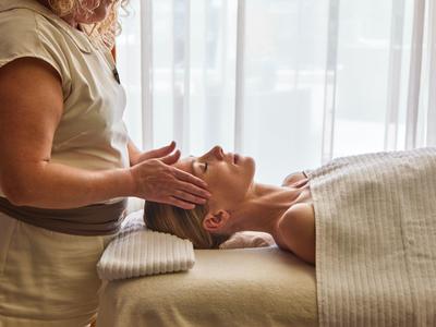 Vrouw krijgt gezichtsbehandeling in lichte, rustige spa-ruimte naast raam met gordijnen.