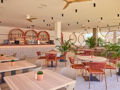 Modern restaurant met houten stoelen, tafels en groene planten in een lichte inrichting.