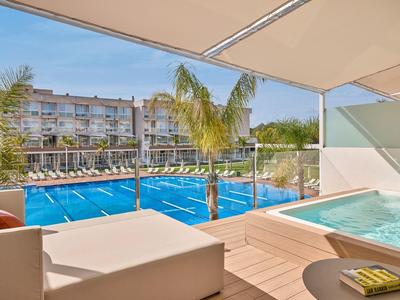 Terras met bubbelbad, ligstoelen en uitzicht op een zwembad en hotelgebouw bij zonnig weer.