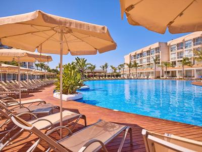 Modern hotelzwembad met ligstoelen en parasols onder heldere blauwe lucht.