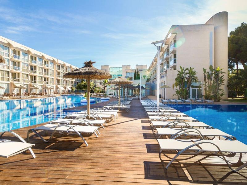 Houten loopbrug tussen twee zwembaden met ligstoelen en moderne hotelgebouwen op de achtergrond