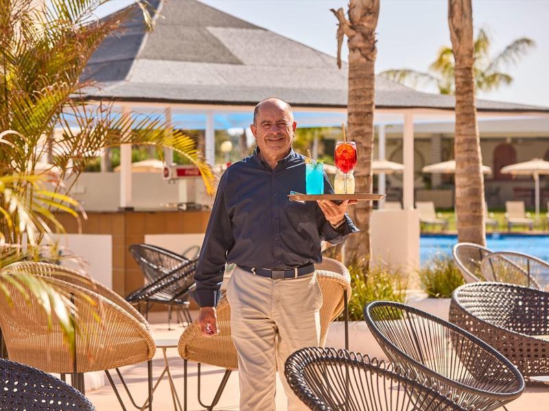 Een ober draagt kleurrijke cocktails in een zonnig zwembadgebied met ligstoelen en palmbomen.