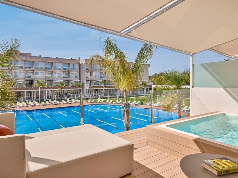 Terras met bubbelbad, ligstoelen en uitzicht op een zwembad en hotelgebouw bij zonnig weer.