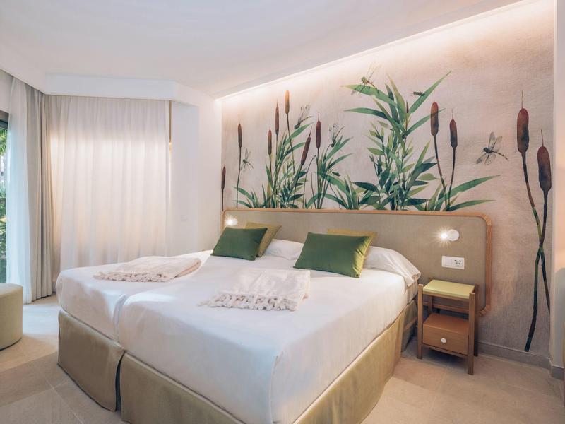 Jasny pokój hotelowy z dwoma łóżkami pojedynczymi i roślinną dekoracją ściany.