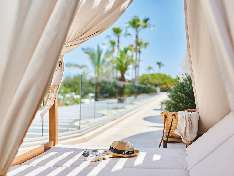Widok przez białe zasłony na strefę basenową z palmami i wygodnymi miejscami do siedzenia.