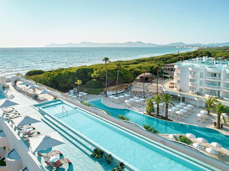 Hotel z długim basenem na zewnątrz, leżakami i widokiem na morze w słoneczny dzień.