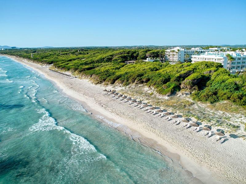 Biała piaszczysta plaża z parasolami i turkusowym morzem przed zielonymi wydmami i hotelami.