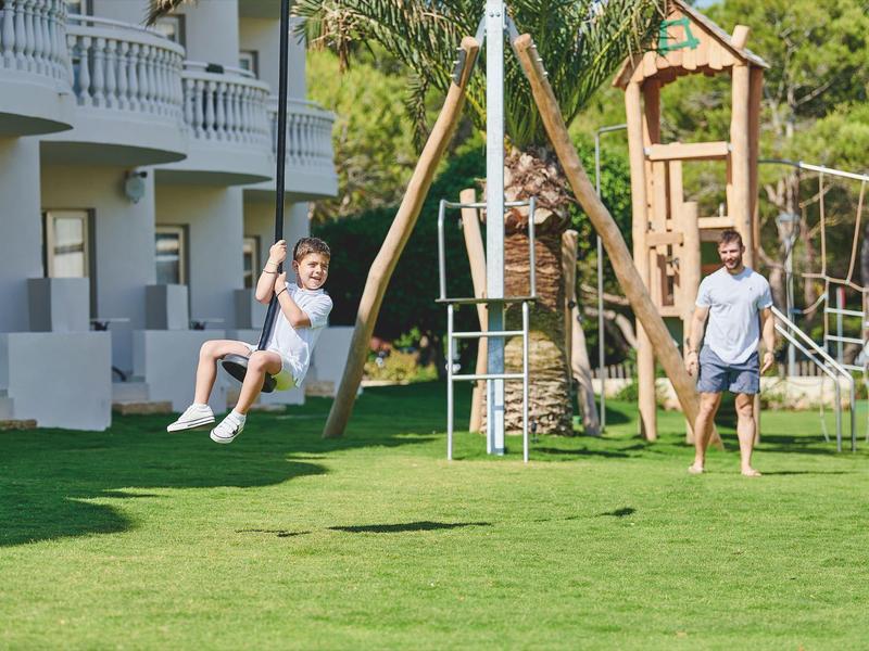 Kobieta huśtająca się w ogrodzie hotelowym, mężczyzna patrzy, plac zabaw w tle