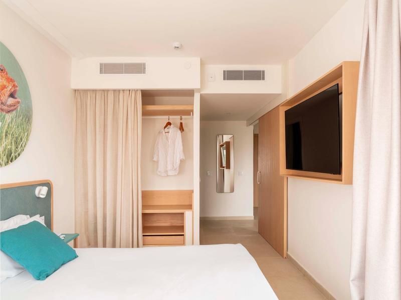 Nowoczesny pokój hotelowy z otwartą szafą, łóżkiem i telewizorem na ścianie.