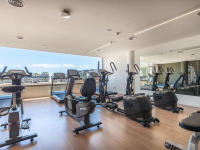 Modernes Fitnessstudio mit Cardiogeräten, Spiegelwand, Holzboden und großen Fenstern mit Aussicht.