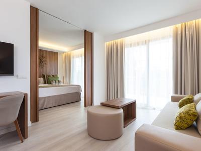 Helles Wohnzimmer mit beigem Sofa, großem Spiegel, Fernseher und hellen Vorhängen an großen Fenstern.
