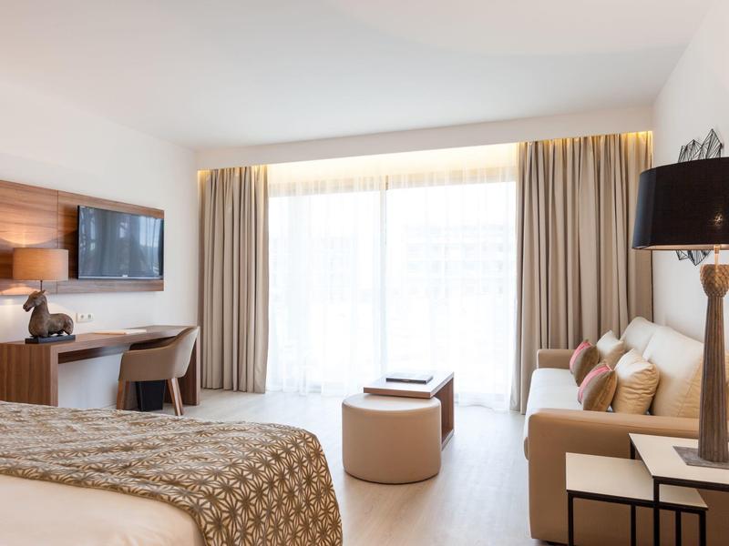 Bright hotel room with bed, desk, sofa, and large window with curtains.