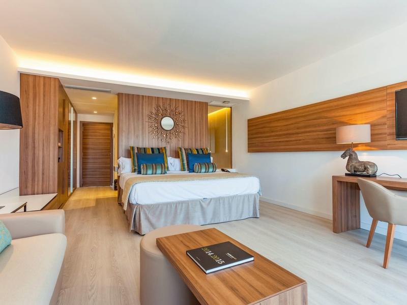 Bright hotel room with double bed, wooden furniture, and seating area with sofa and table.
