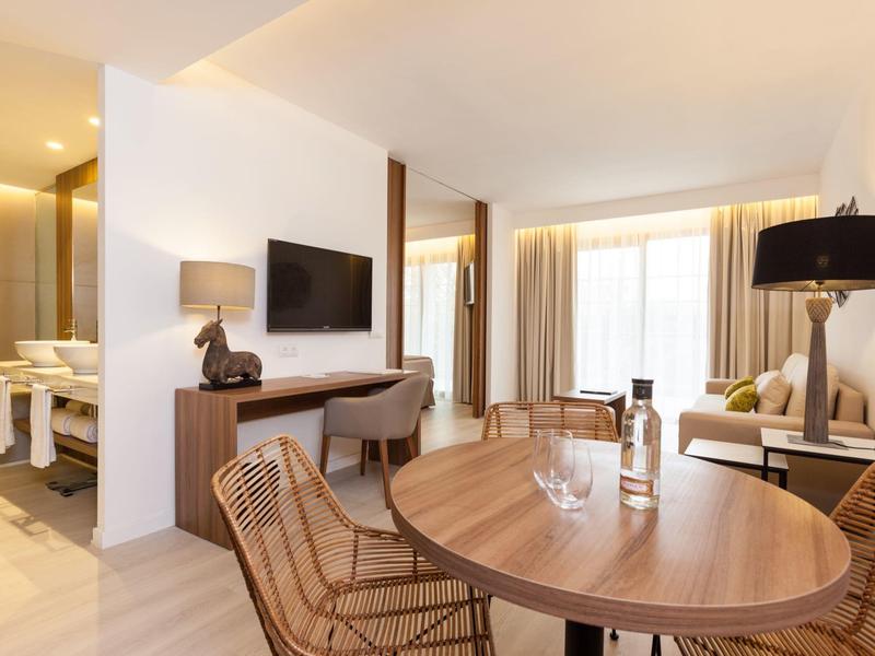 Bright hotel room with dining area, sofa, desk, and modern bathroom in the background.