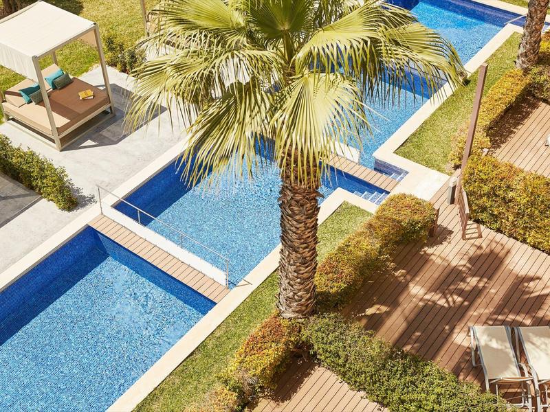 Pool area with palm trees, sun loungers, and a covered daybed in a hotel complex