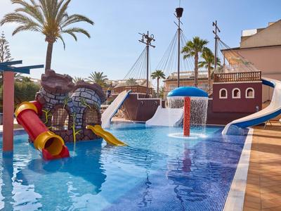 Children's pool with water slides and water features next to palm trees and buildings on a sunny day.