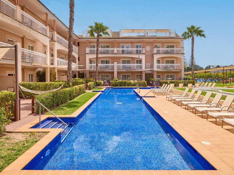 Modern outdoor pool with lounge chairs and palm trees in front of a hotel building under clear sky.