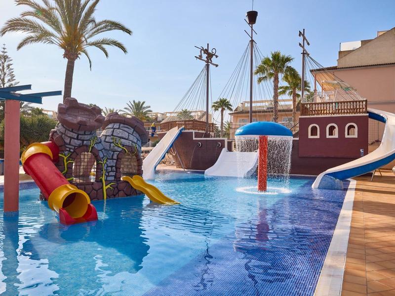 Children's pool with water slides and water features next to palm trees and buildings on a sunny day.