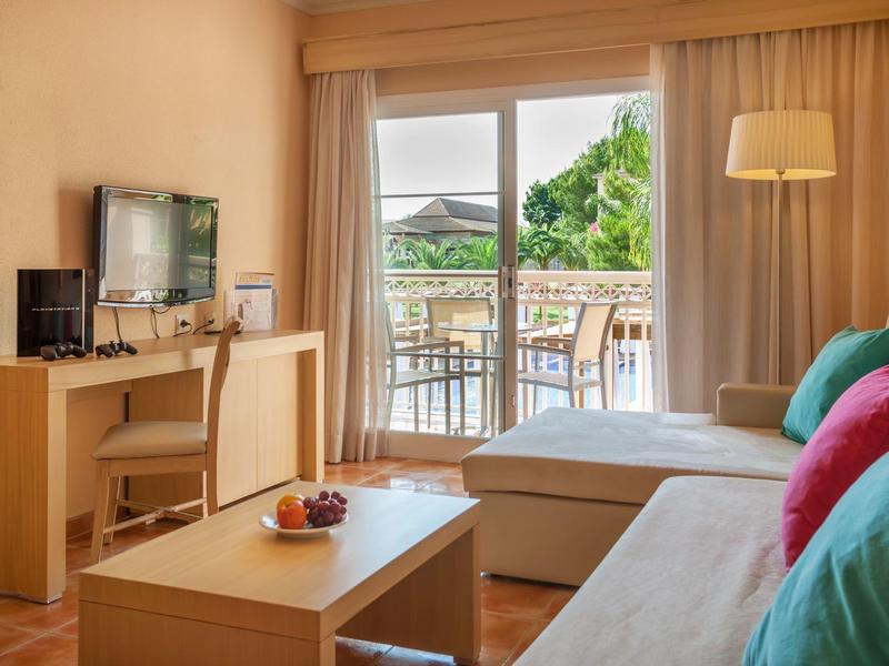 Bright living room with sofa, colorful cushions, TV, desk, and balcony with green view.