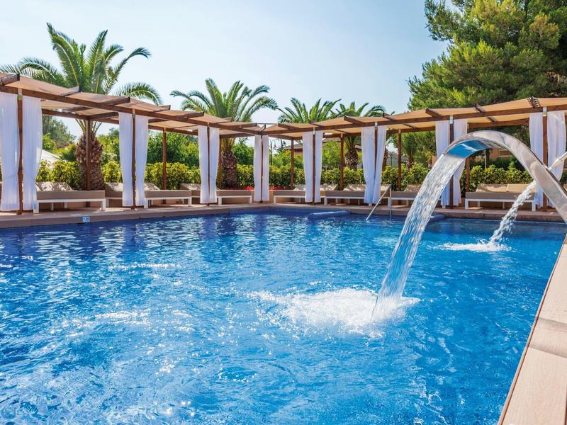 Modern pool with waterfall and covered seating areas surrounded by palm trees.