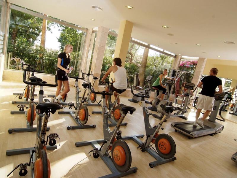 Gym with stationary bikes and treadmills, three people exercising, large windows with garden view.