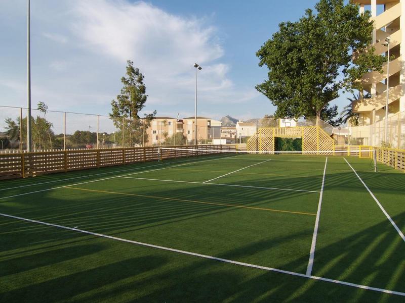 A tennis court with green artificial turf and white lines next to a building and trees.