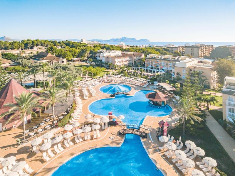 Large hotel pool with sun umbrellas and loungers, surrounded by hotel buildings and green landscape.