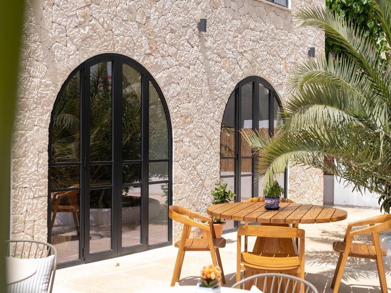 Terrace with round wooden tables and chairs in front of arched windows of a stone house.