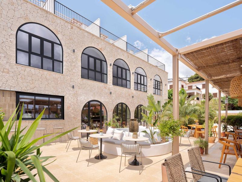 Sunny terrace with seating in front of a hotel featuring large arched windows.