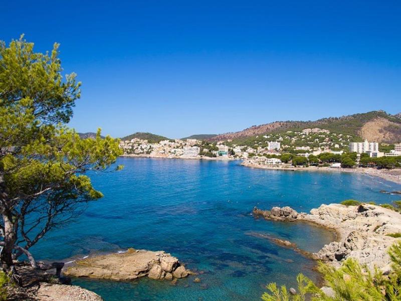 Clear blue sea with rocks, trees, and a coastal town under a bright blue sky.
