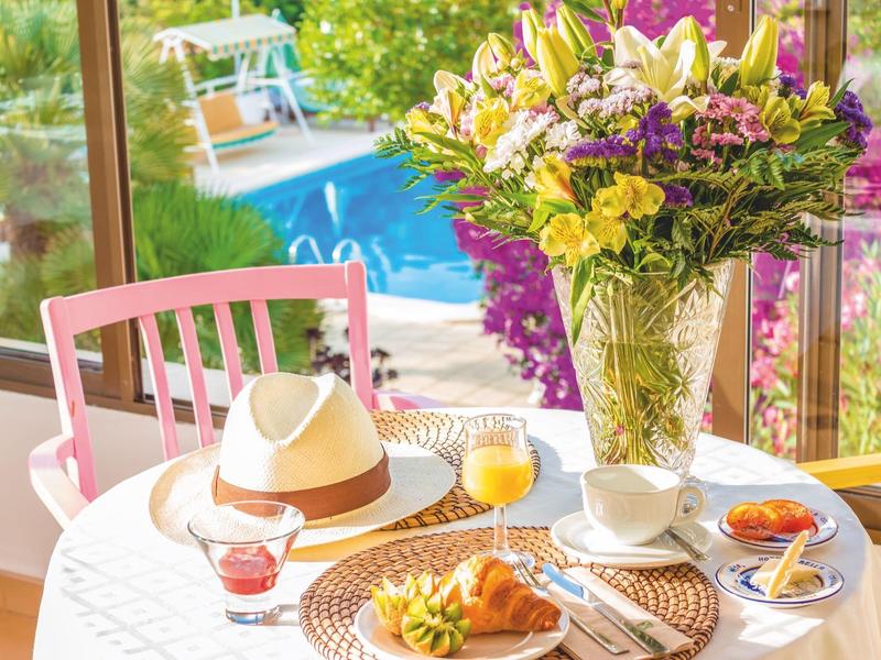 Breakfast table with pastries, juice, and flowers by a panoramic window overlooking a pool.