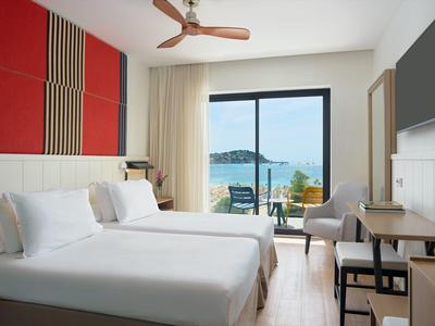 Lichte hotelkamer met twee bedden, ventilator, zithoek en zeezicht vanaf het balkon.