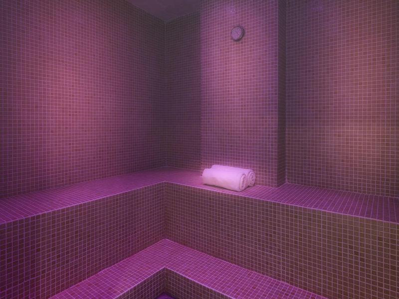 Interieur van een kleine sauna met roodachtig verlichte mozaïektegels en handdoeken.