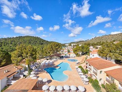 Großer Hotelpool mit Sonnenschirmen, umgeben von Bäumen und Bergen unter blauem Himmel.