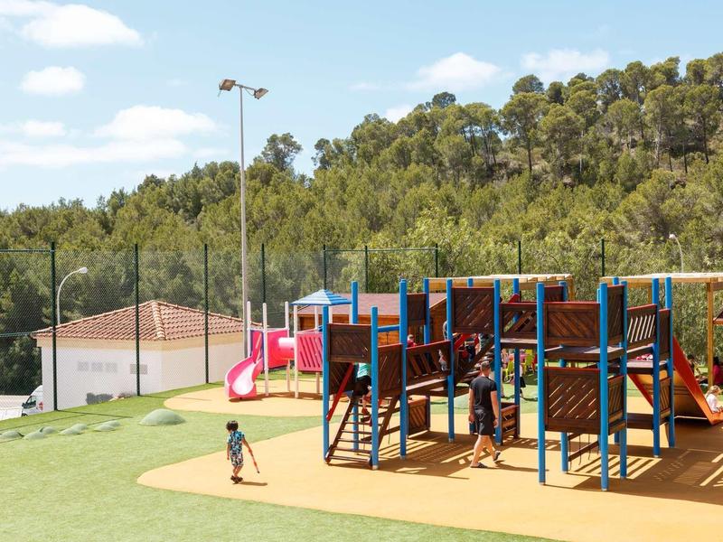 Kinderspielplatz mit Klettergerüst, Rutsche und Grasfläche vor bewaldetem Hügel.