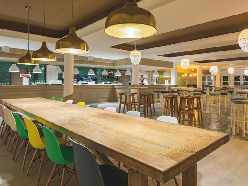 Großer Gemeinschaftstisch mit bunten Stühlen, Holzvertäfelung, modernen Lampen und Barbereich.