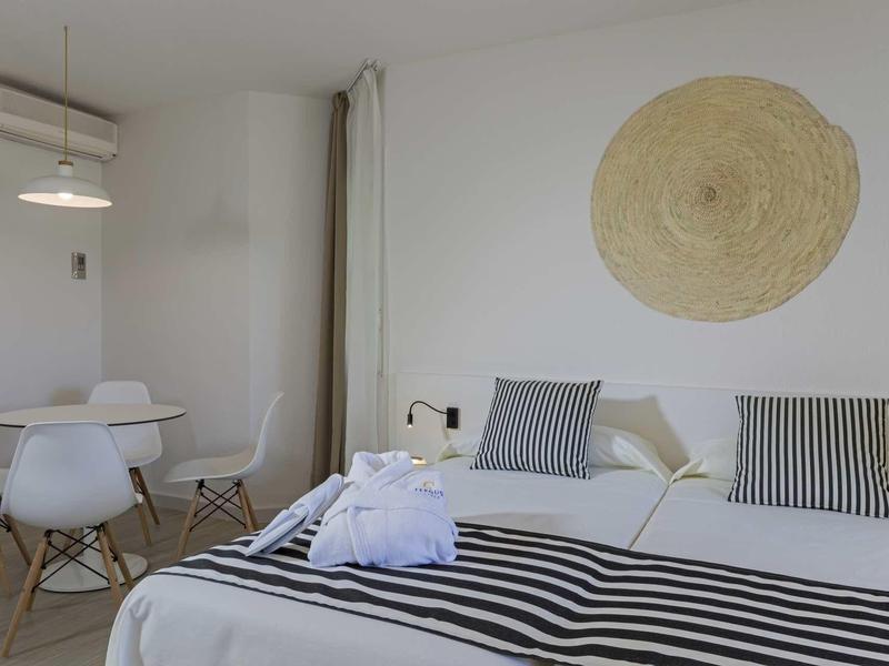 Helles Schlafzimmer mit weißem Bett, gestreiften Kissen, Wanddeko und kleinem Essbereich.