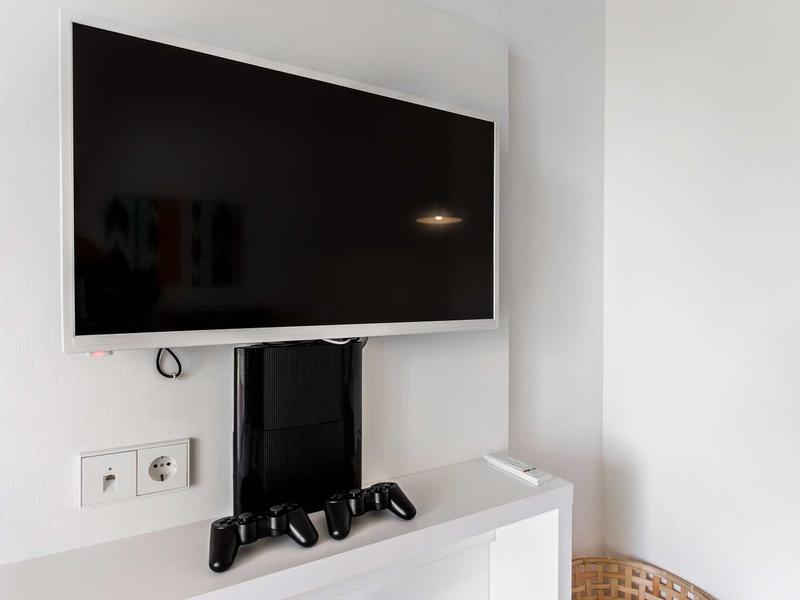 Großer schwarzer Fernseher hängt an weißer Wand über weißem Möbelstück mit zwei Spielcontrollern.