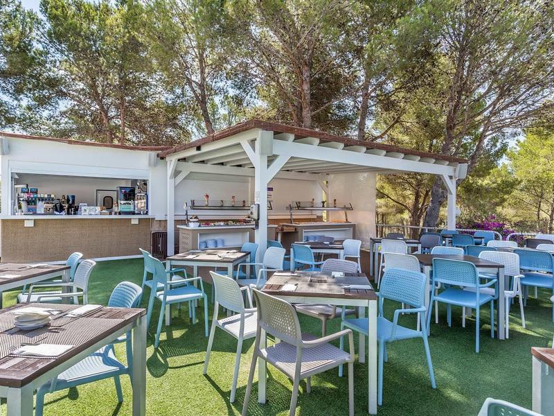 Außeriridischer Sitzbereich mit blauen Stühlen, Holztischen und weißem Holzpavillon unter Bäumen.