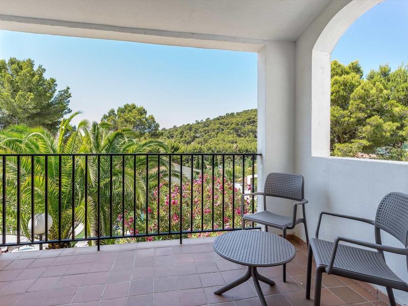 Balkon mit zwei Stühlen, kleinem Tisch, weißem Geländer und Blick auf grüne Bäume und Hügel.